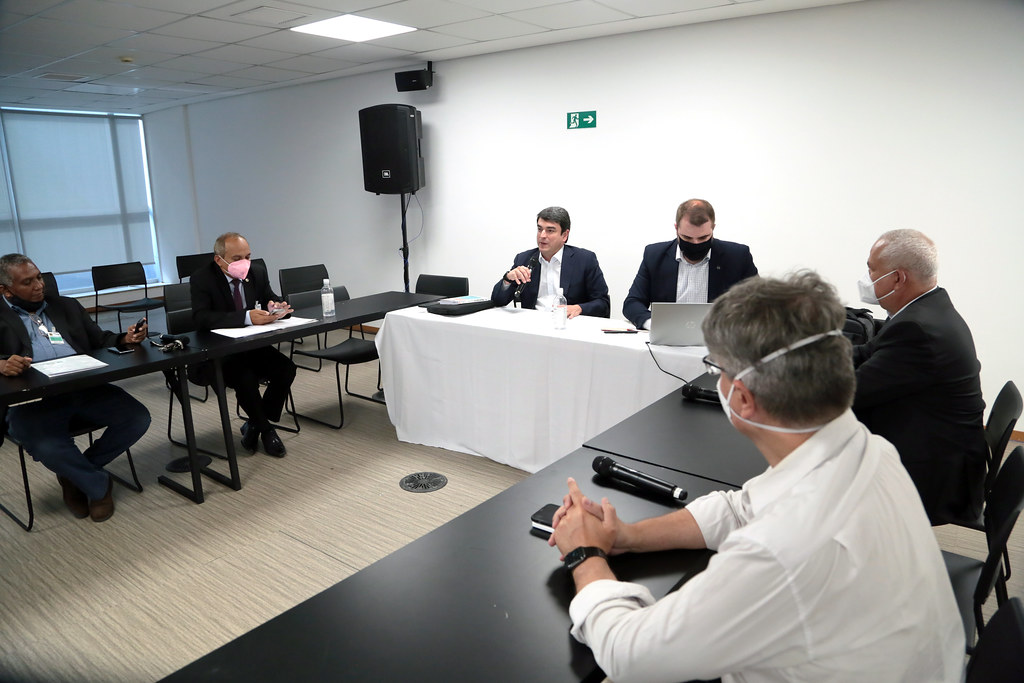 Foto mostra pessoas participantes presencialmente na reunião, sentados em mesa em U.