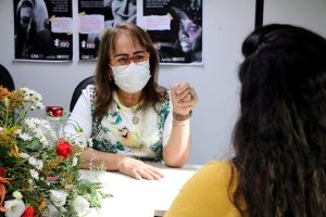 Foto da juíza Hermínia Azoury falando com uma mulher, ambas sentadas em uma mesa de trabalho.