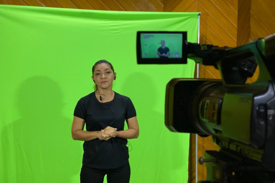 Foto mostra uma mulher vestida de calça e camiseta pretas à frente de uma tela de verde para chroma-key. Ela está posicionada em frente a um a filmadora com tripé, onde é possível ver a imagem da mulher no visor.