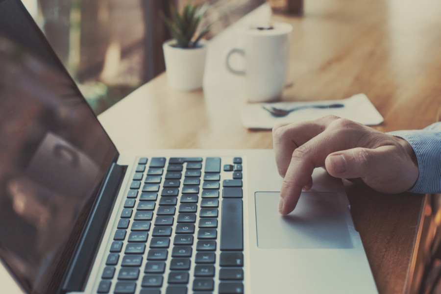 Foto mostra a mão de um homem usando um notebook sobre uma mesa de trabalho.