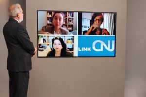 Foto mostra apresentador do programa, no estúdio, olhando para o telão onde se vê, por videoconferência, as três entrevistadas e a logomarca do programa Link CNJ.