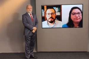 Foto mostra o apresentador do programa no estúdio, ao lado do telão onde se vê as duas pessoas entrevistadas.