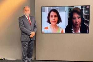 Foto mostra o apresentador do programa no estúdio, posando ao lado do telão onde se vê as duas entrevistadas por videoconferência.