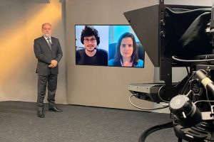 Foto no estúdio de gravaçlão do programa mostra o apresentador posando ao lado de telão onde se vê as duas pessoas entrevistadas.