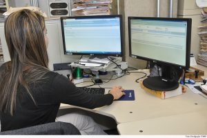 Foto mostra uma mulher, sentada em uma estação de trabalho, usando o computador com dois monitores, onde acessa o PJe.