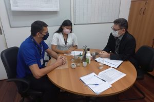 Foto mostra dois homens e uma mulher conversando em uma sala, sentados em volta de uma mesa redonda, com papeis na frente deles.