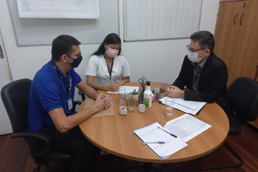 Foto mostra dois homens e uma mulher conversando em uma sala, sentados em volta de uma mesa redonda, com papeis na frente deles.