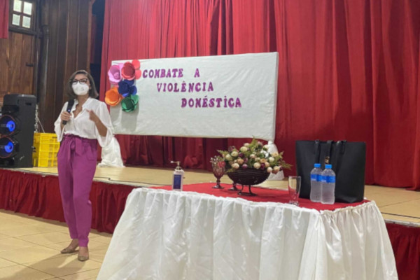 Foto mostra momento de evento realizado durante a 20ª Semana Justiça Pela Paz em Casa, com uma mulher no palco e, ao fundo, um cartaz onde se lê "Combate à Violência Doméstica"