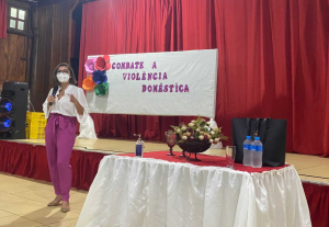 Foto mostra momento de evento realizado durante a 20ª Semana Justiça Pela Paz em Casa, com uma mulher no palco e, ao fundo, um cartaz onde se lê "Combate à Violência Doméstica"