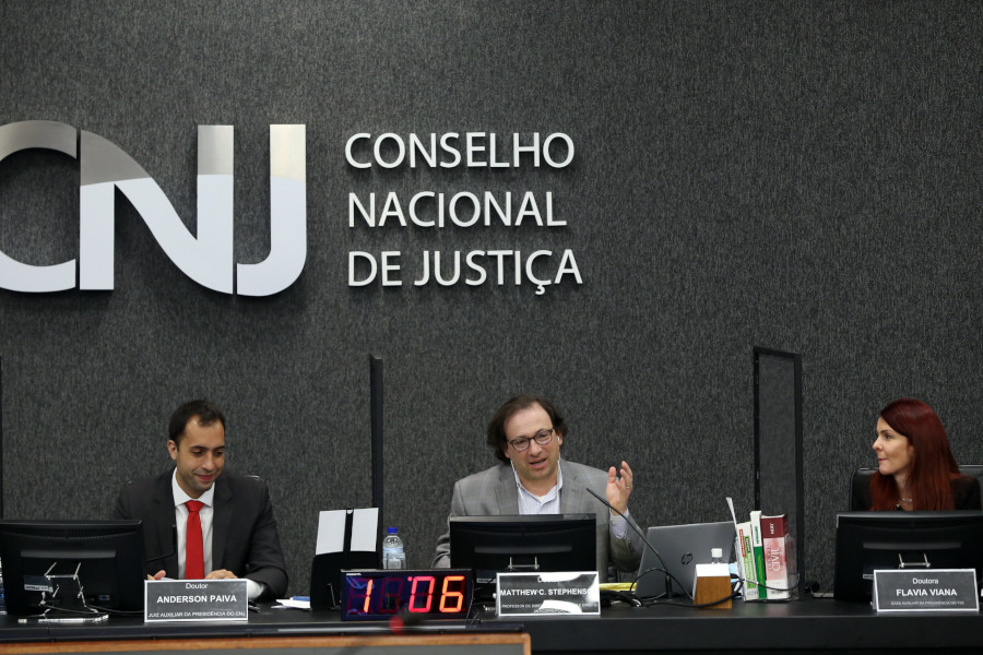 Foto mostra a bancada do plenário do CNJ durante o evento, com o juiz auxiliar da Presidência do CNJ Anderson de Paiva à esquerda; o professor da Harvard Law School Matthew C. Stephenson ao centro; e a juíza auxiliar da Presidência do TSE Flávia da Costa Viana à direita.