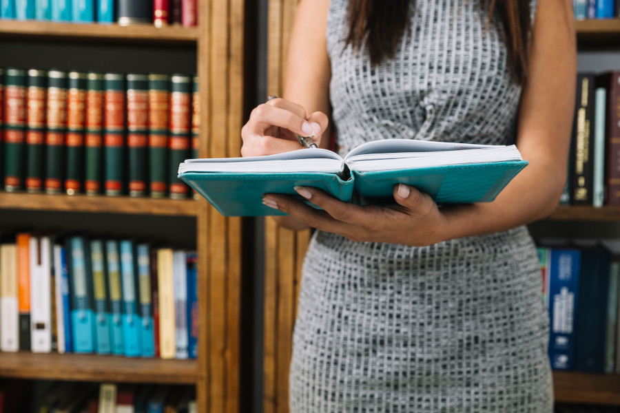 Foto mostra uma mulher em pé, segurando um livro e fazendo anotações nele com uma caneta. Ela está em um ambiente onde há diversas estantes com livros atrás dela.