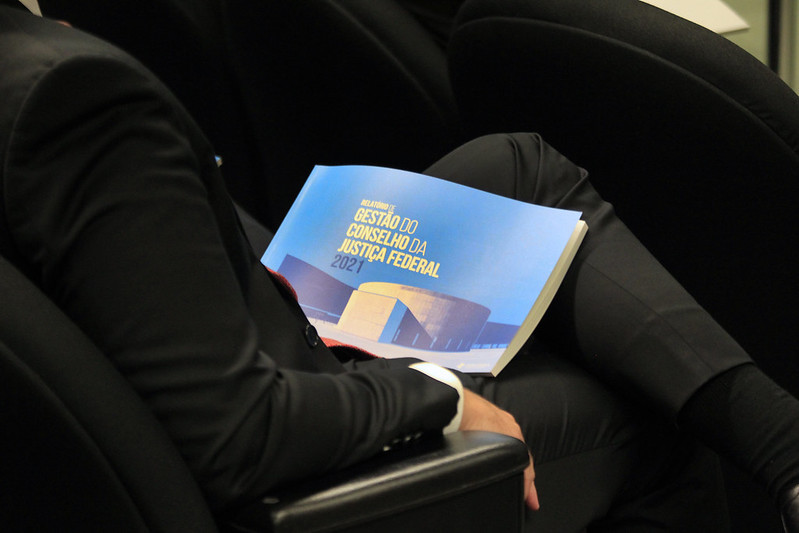 Foto mostra um homem de terno sentado com um relatório impresso no colo.