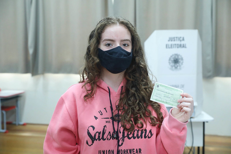 Foto mostra, em uma seção eleitoral, uma jovem usando máscara de proteção contra a Covid- 19 e mostrando seu o título de eleitor.