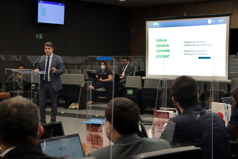 Foto mostra uma sala, com um telão ao fundo. Um pouco ` a frente e à direita do telão, o conselheiro Bandeira de Mello está falando para um grupo de pessoas sentadas.