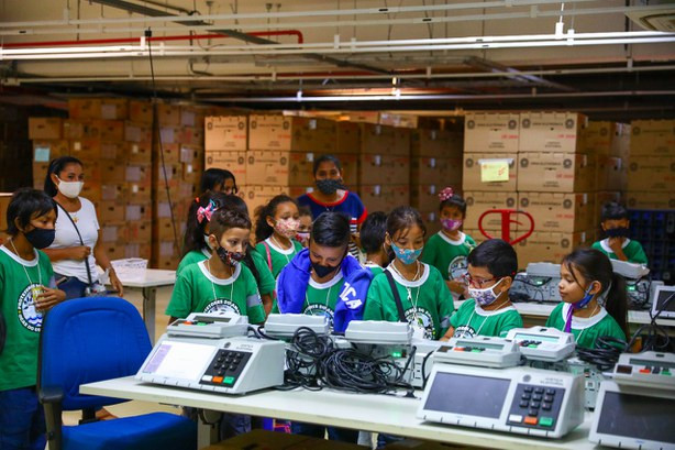 Foto mostra crianças de escola municipal visitando um espaço onde há vários modelos de urnas eletrônicas.