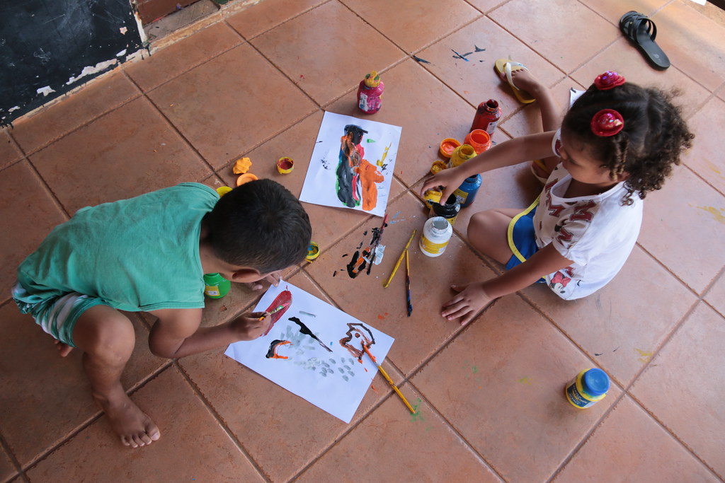 Foto mostra duas crianças desenhando sentadas no chão. Há diversas tintas, pinceis e folhas com os desenhos.