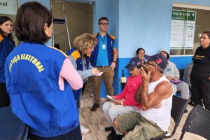 Foto mostra pessoas sentadas em cadeiras aguardando atendimento enquanto equipe do TRE-PR presta algumas informações.