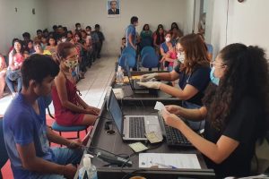 Foto mostra uma sala, com várias sentadas ao fundo aguardando para serem atendidas e, à frente, balcão de atendimento, com duas atendentes sentadas e atendendo a uma mulher e a um jovem.