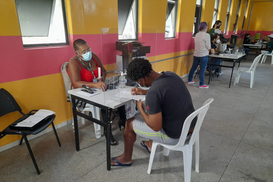 Foto mostra um homem sentado em uma cadeira escrevendo em uma folha. Ele está sendo atendido por uma sentada do outro lado da mesa.