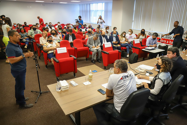 Foto mostra momento de audiência pública em Governador Valadares, com uma pessoa falando ao microfone enquanto se vê um auditório cheio e sentados, na bancada, o conselheiro Bandeira e representantes do MPMG.