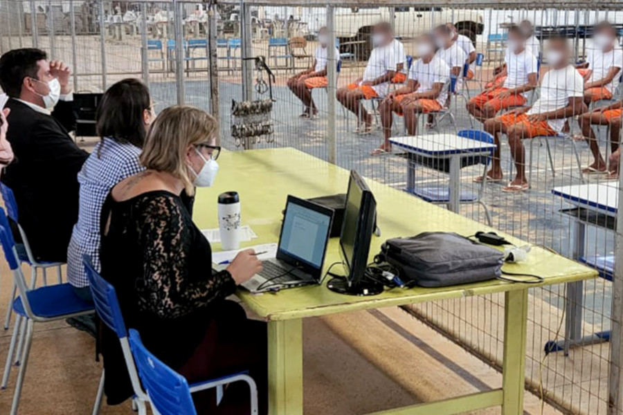 Foto mostra de audiência com pessoas presas em penitenciárias, sendo visível à esquerda as representantes do Judiciário, sentadas atrás de uma mesa, e as pessoas internas, todas de uniforma, sentadas em cadeiras à direita. Entre os dois grupos, há uma grade divisora.