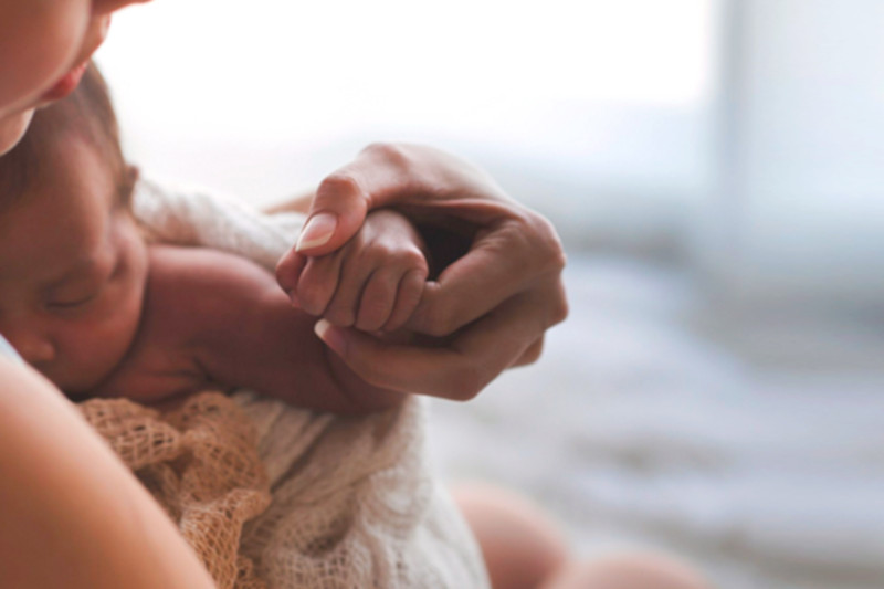 Foto mostra a mão de uma mulher adulta segurando a mão de um bebê que está em seu colo.