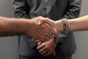 Foto mostra as mãos de um homem de uma mulher se cumprimentando, ` à frente de um homem usando terno e gravata.