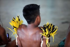 Foto das costas de criança indígena sentada no chão, sem camisa e usando um adereço indígena nos ombros.