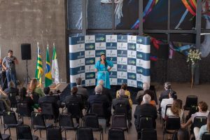 Foto mostra visão geral do evento de instalação, com a ouvidora nacional da Mulher, Tânia Reckziegel, falando em pé em frente a um backdrop com a marca do TRT18 e as pessoas à sua frente, sentadas e assistindo.