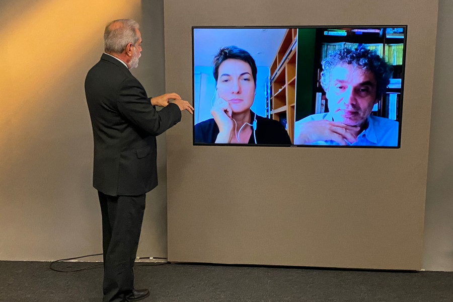 Foto mostra o apresentador do programa no estúdio, virado para o telão na parede e conversando com os dois entrevistados por videoconferência.