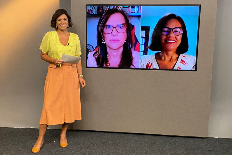 Foto mostra, no estúdio, a apresentadora do programa sorrindo ao lado do telão onde se vê as duas entrevistadas por videoconferência.