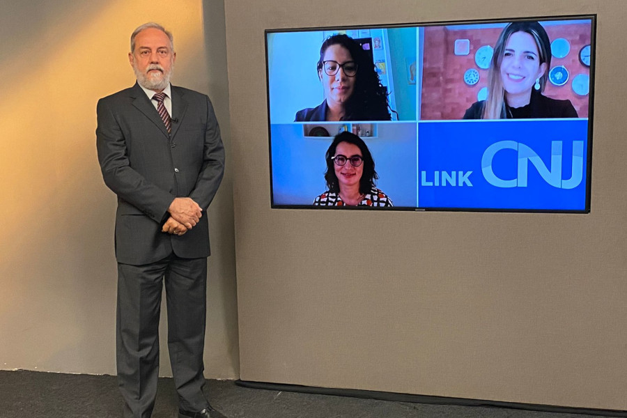 Foto no estúdio do programa mostra o apresentador posando em pé ao lado da telão, onde estão os rostos das três entrevistadas da edição.