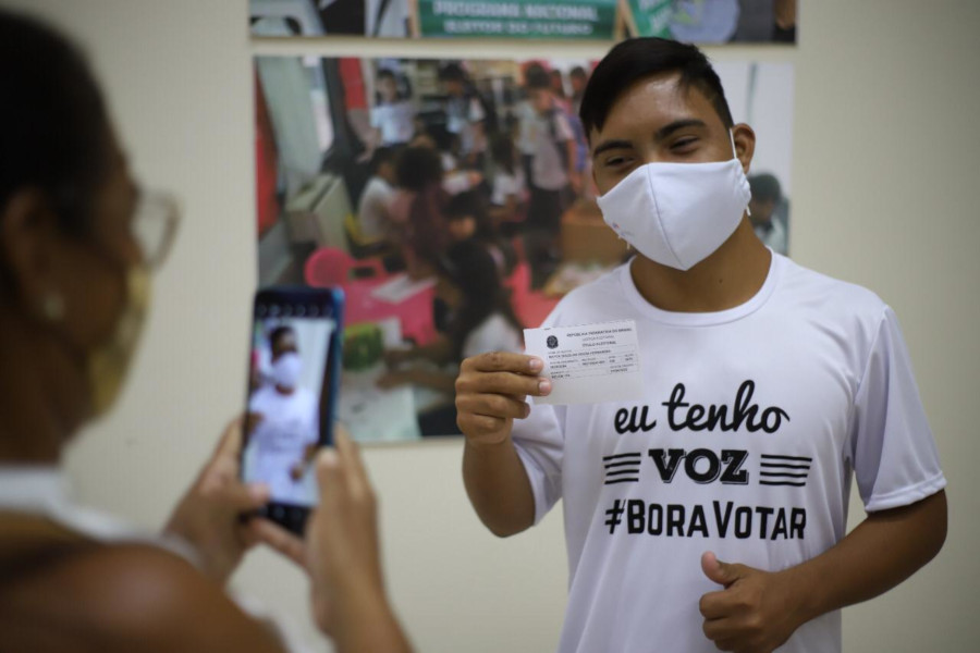 Foto de homem com síndrome de down segurando recibo de regularização eleitoral. Ele está posando para um outras pessoa, que está o fotografando com um celular.