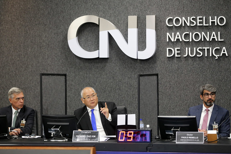 Foto mostra o conselheiro do CNJ Pae Kim falando durante a reunião no Plenário do CNJ. Ele está sentado na bancada principal, ao lado de dois outros membros do Fórum.
