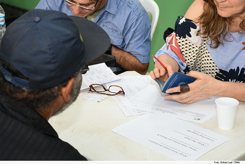 Foto mostra um homem sentado e sendo atendido por uma mulher que segura uma carteira de trabalho.