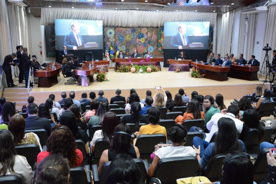 Foto mostra visão geral do auditório do TJRR, com diversas pessoas sentadas assistindo. No palco, tem uma bancada com representantes de instituições e dois grandes telões onde aparece o palestrante dos EUA falando.