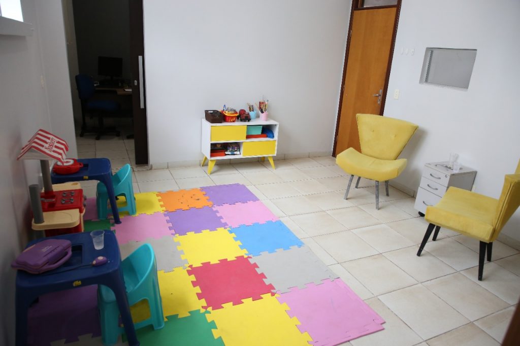 Foto mostra sala, com diversos brinquedos, tapete para brincar, duas mesas e cadeiras para crianças sentarem e poderem desenhar, além de duas poltronas.