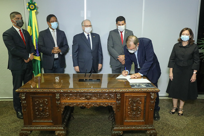 Foto mostra, atrás de uma mesa de madeira na sede do TSE, quatro homens e uma mulher em pé, ao fundo, enquanto um outro homem assina o acordo na mesa.