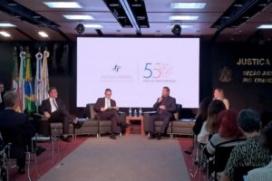 A foto mostra pessoas sentadas em cadeiras no auditório. Na frente, há dois sofás de dois lugares cada um posicionados próximos e de frente para a plateia. Neles estão sentados 3 homens, vestindo terno, e uma mulher. Ao fundo, o telão do evento com o logo da justiça federal e do evento: 55 anos de transformação