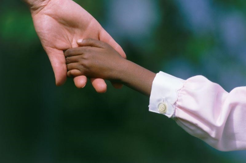 Foto mostra a mão de um homem branco segurando a mão de uma criança negra.