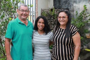 Foto mostra a adolescente abraçada e sorrindo com pai e mãe adotiva.