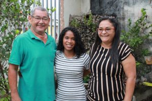 Foto mostra a adolescente abraçada e sorrindo com pai e mãe adotiva.