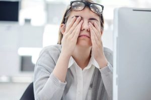 Foto mostra uma mulher, sentada em uma estação de trabalho e com os óculos em cima da cabeça, enquanto coça os olhos como se estivesse cansada.