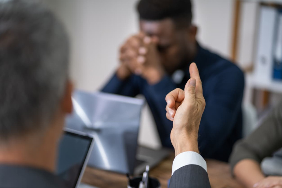 Foto mostra dois homens sentados em uma mesa. Ao fundo, um homem negro está com o rosto entre as mãos, cabisbaixo. À sua frente, de costas, um homem mais velho apontando o dedo de forma acusadora.