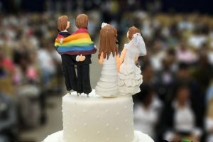 Foto mostra em destaque a parte de cima de um bolo de casamento, onde estão bonecos de um casal de homens de terno enrolados em uma bandeira do arco-íris e um casalo de mulheres vestidas de noiva.