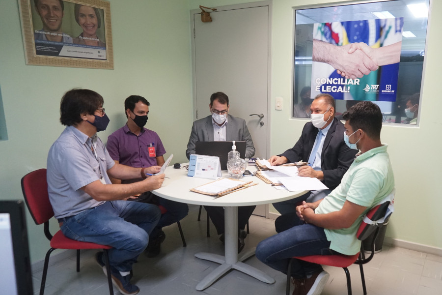 Foto mostra cinco homens sentados em volta de uma mesa redonda em uma sala, onde estão buscando acordo em um processo. Na parede da sala tem um cartaz com duas mãos se cumprimentando e o texto "Conciliar É Legal!"