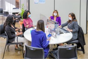 Foto mostra mesa de conciliação em uma sala. São cinco mulheres sentadas em volta de uma mesa, todas usando máscaras protetoras contra a Covid-19. No meio da mesa, tem uma tela de acrílico, também para proteção.