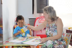 Foto mostra Constância fazendo um carinho em João Miguel. Ambos estão sorrindo em uma sala com brinquedos.