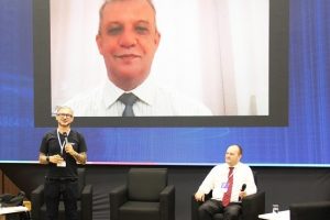 Foto mostra um palco grande. À frente, está um homem em pé com um microfone. Próximo a ele, um outro homem está sentado em uma poltrona. E, ao fundo deles, em um grande telão, tem um terceiro homem participando por videoconferência.