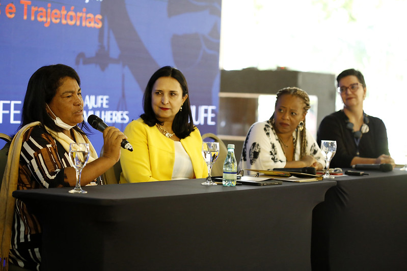 Foto mostra as quatro mulheres participantes do painel sentadas atrás de mesa no palco. No primeiro plano, Rute Fiúza está com o microfone, fazendo sua fala, quanto as demais olham para ela.